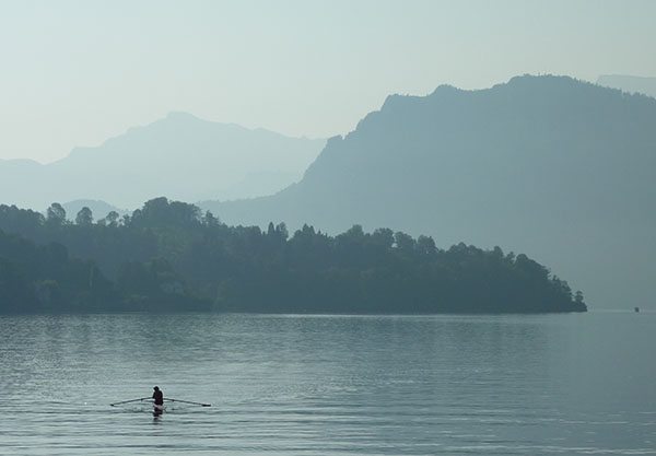 A person rowing on his boat during an early morning scene in Lake Lucerne. The background with the trees and mountains are a bit foggy, with the farthest one slowly fading.